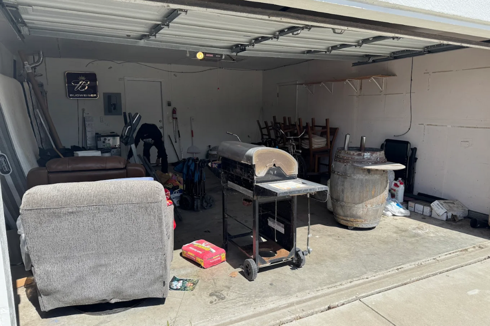A cluttered residential garage in Fairfield filled with old furniture, a grill, and an oak barrel before a cleanout service.